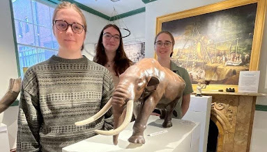 Three women stand behind a scale model of a wooly mammoth