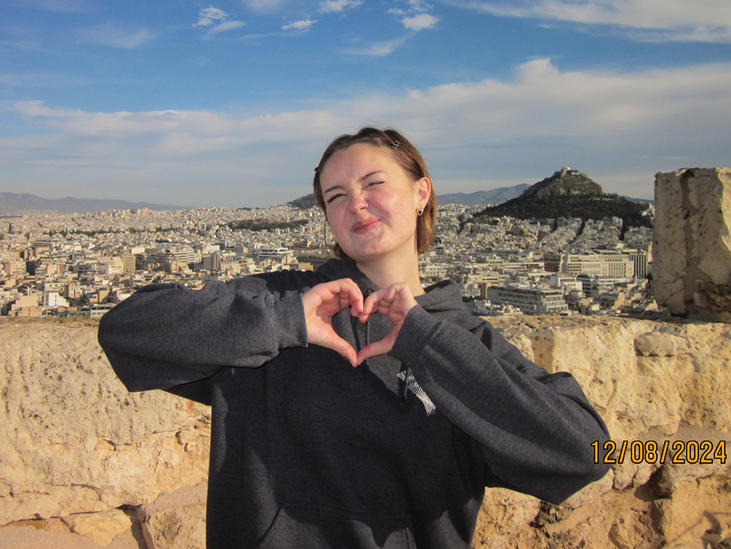 Eva Burcher stands in front of a city backdrop, making a heart symbol with her hands