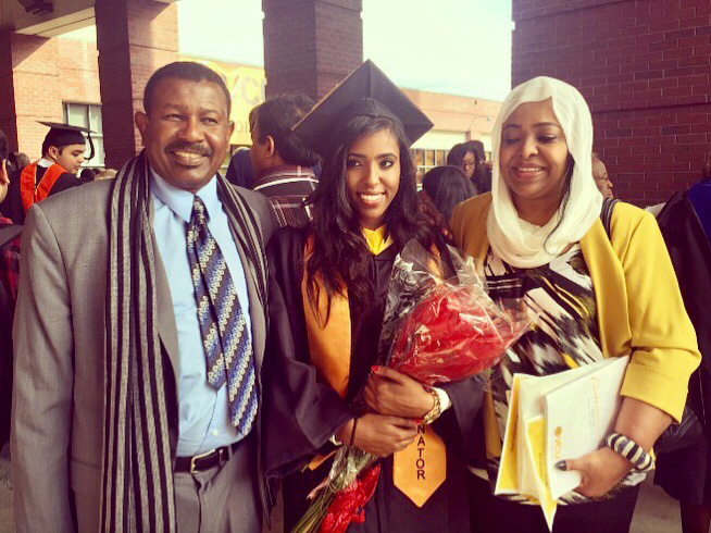 Isra'a Nour in a black cap and gown with a gold sash, holding a wrapped bouquet of red flowers. She stands between an older man in a grey suit and striped scarf, and an older woman in a yellow cardigan and white headscarf.