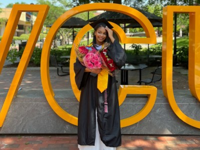 VCU International Studies alum Ithar Hassan (Class of 2023) wears a cap and robe and holds flower bouquets in front of a VCU sign on the Monroe Park campus.